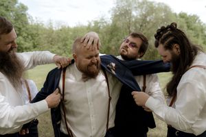 Groom being playfully helped into jacket by groomsmen and bridesmaid in outdoor wooded setting.