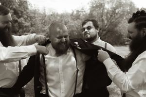 Black and white photo of groomsmen helping the groom adjust his suit jacket outdoors before a wedding ceremony.