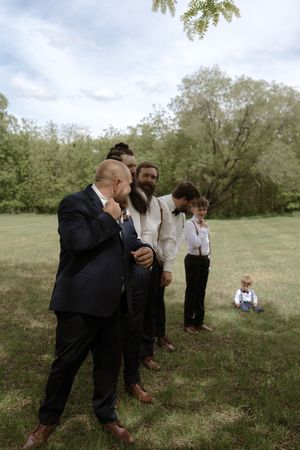 Groom in navy suit stands with groomsmen in suspenders on green lawn before ceremony.