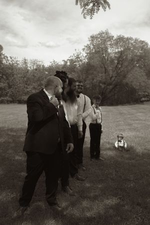 Black and white photo of groomsmen laughing together on a lawn with a small child sitting in the background.