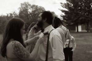 Black and white photo of tattooed woman adjusting groom's boutonniere outdoors before wedding ceremony.