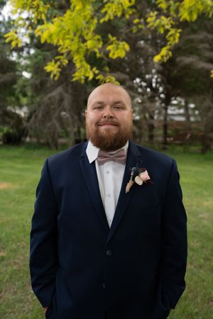 Bald groom with beard smiles in navy suit with pink bow tie and boutonniere outdoors.