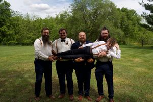 Groomsmen in navy suits playfully lift groom in white shirt outdoors on green lawn.