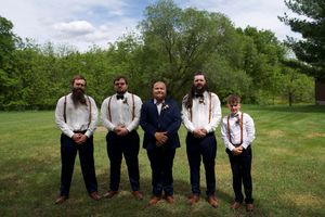 Five groomsmen in white shirts, suspenders, and dark pants posing together on a green lawn at a wedding.