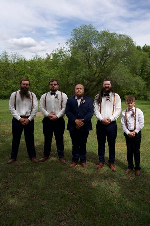 Groom in navy suit stands with four groomsmen in white shirts and suspenders on a grassy field.