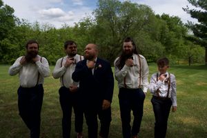 Groom with groomsmen in white shirts and suspenders laughing together on green lawn before wedding ceremony.