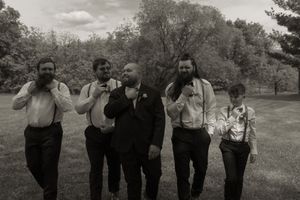 Black and white photo of groomsmen in suspenders and bow ties laughing outdoors at a wedding.