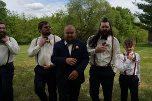 Groom in navy suit stands with groomsmen wearing suspenders on green lawn at wedding.