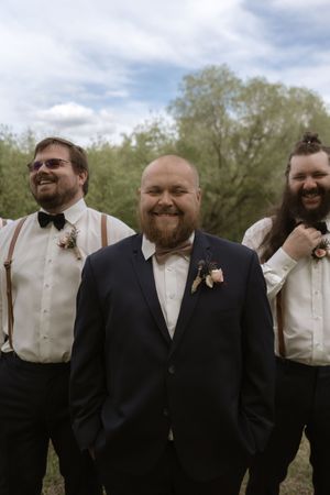 Groom in navy suit with bow tie stands with groomsmen in suspenders and bow ties outdoors.