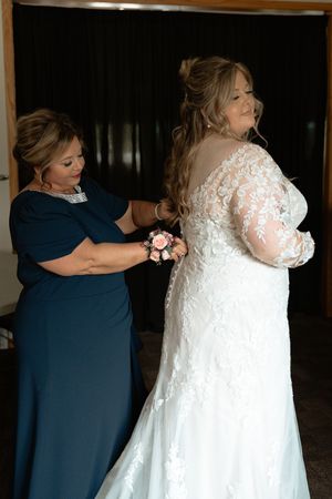 Mother in navy dress helps bride in lace long-sleeve gown with dress buttons before wedding ceremony.