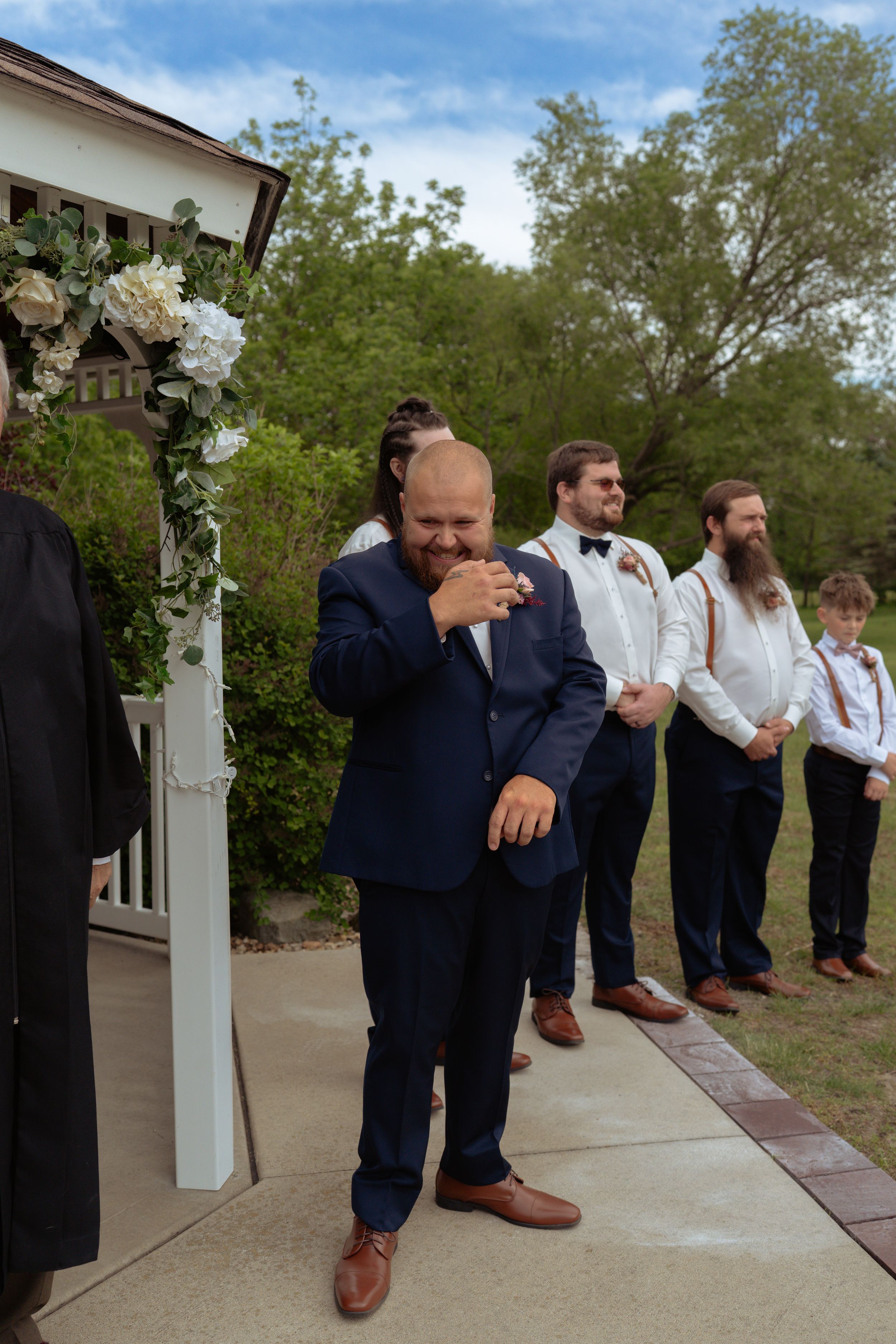 Tearful groom in navy suit reacts emotionally seeing bride walk down the aisle outdoors.