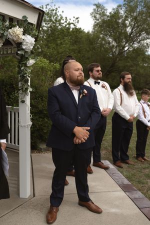 Groom in navy suit with floral boutonniere stands at altar with groomsmen during outdoor wedding ceremony.