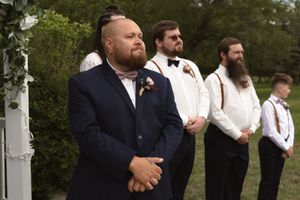 Groom in navy suit standing at altar with groomsmen in white shirts and suspenders during outdoor wedding ceremony.