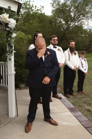 Emotional groom covering face at altar with groomsmen standing behind him outdoors.