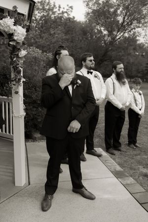 Emotional groom covers face while groomsmen stand behind him at outdoor wedding altar.