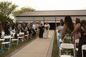 Wedding processional with bride walking down outdoor aisle lined with white chairs and guests near a barn venue.