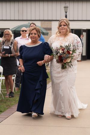Bride in lace gown holding floral bouquet walking down aisle with woman in navy dress at outdoor wedding.