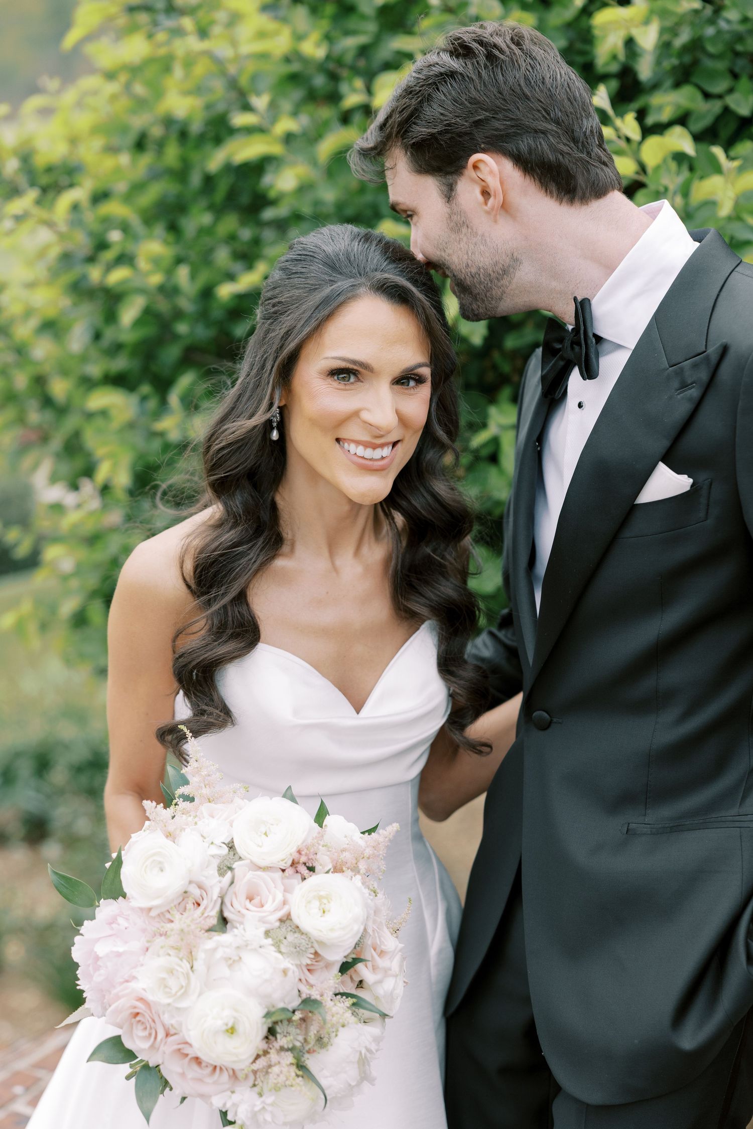 Bride in strapless gown holding blush bouquet smiles as groom in black tuxedo whispers in her ear outdoors.