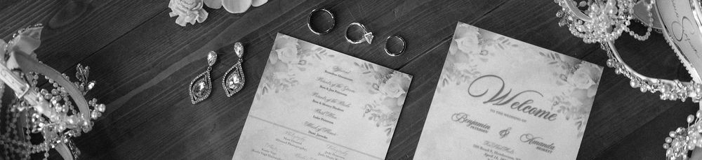 Black and white flat lay of elegant wedding stationery, rings, and pearl jewelry on a wooden surface.