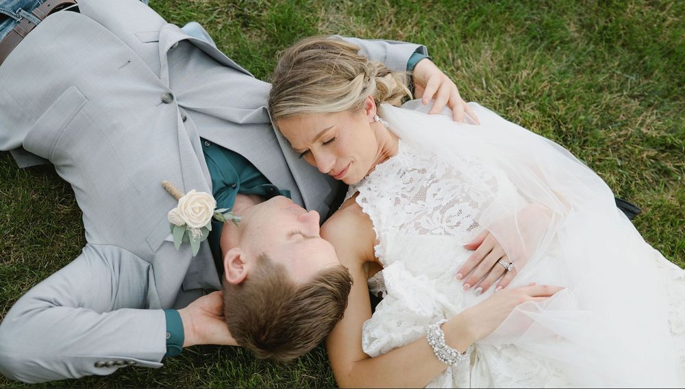 Bride and groom lying on green grass, gazing at each other lovingly in wedding attire, aerial view.