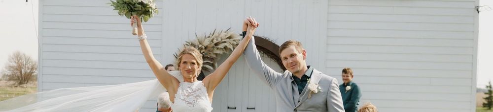 Joyful newlywed couple raising hands in celebration outside a white barn wedding venue.