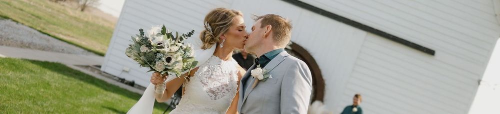 A newlywed couple shares a romantic kiss outdoors near a white barn on their wedding day.