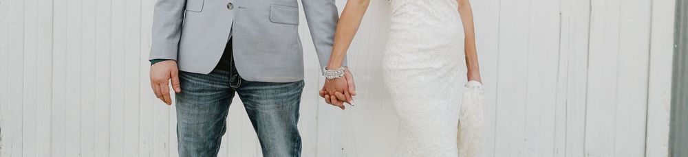 A bride and groom hold hands in front of a white wall, wearing a white wedding dress and light gray suit.