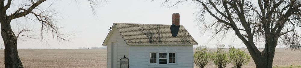 A small white farmhouse with a red chimney stands alone on a flat rural plain, flanked by bare trees.