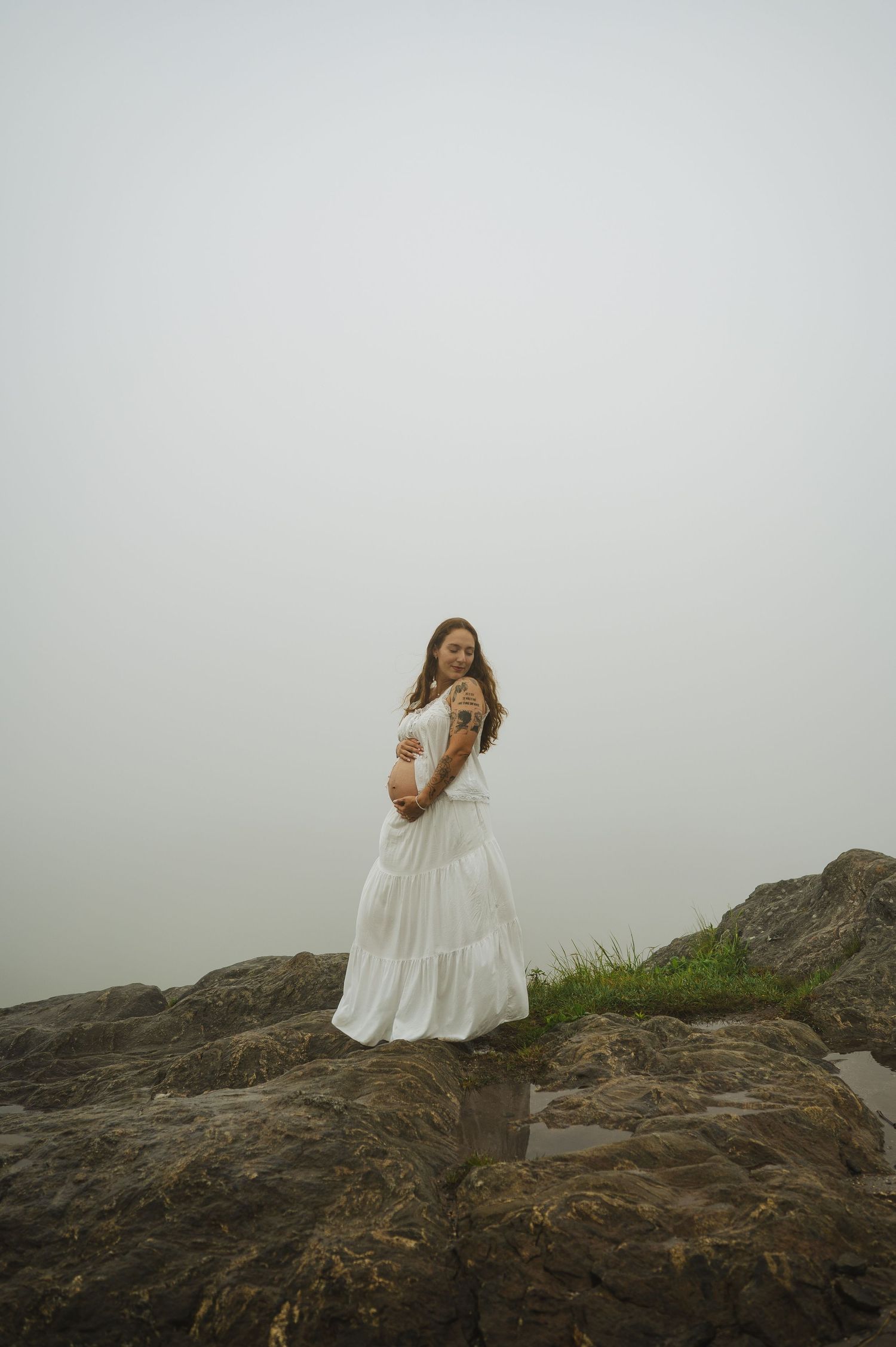 Pregnant woman in white dress standing on rocky coastal cliffs in foggy weather, cradling her baby bump.