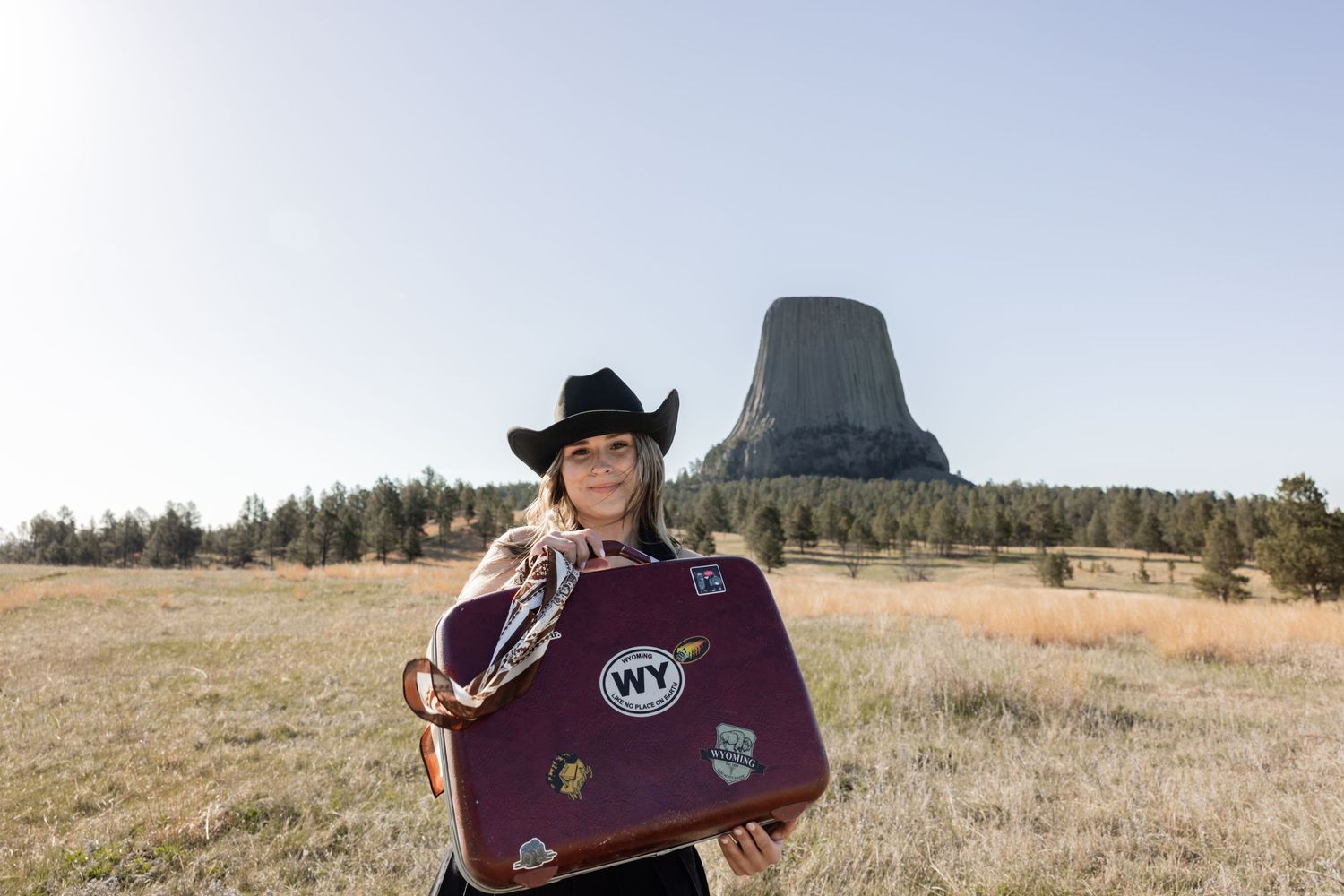 Woman in black cowboy hat holds vintage WY sticker suitcase before Devils Tower, Wyoming landmark.