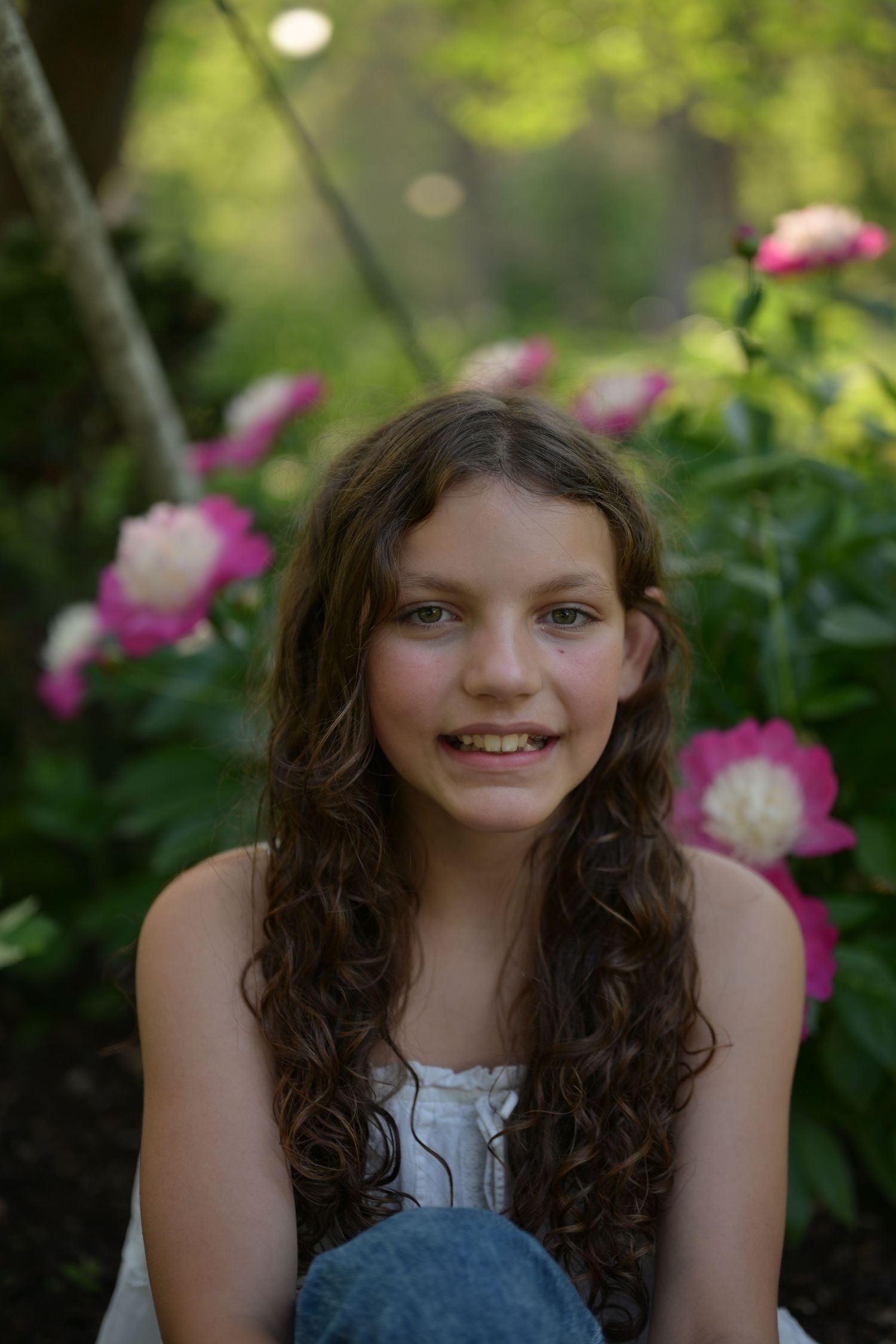 Smiling older woman with white curly hair wearing a white button-up shirt outdoors in natural green setting.