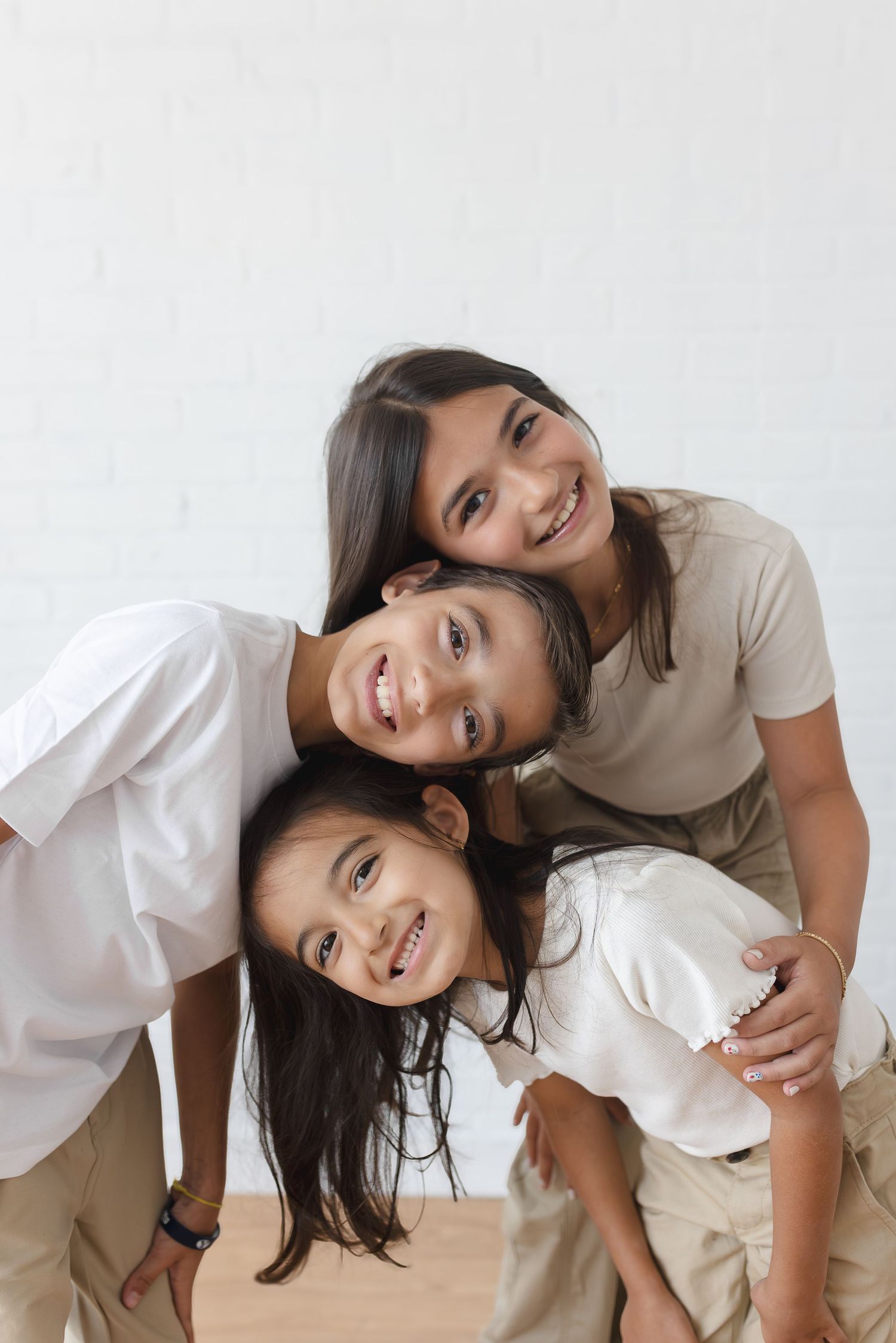 Three smiling girls posing together indoors, wearing casual neutral-toned outfits against a white brick wall.