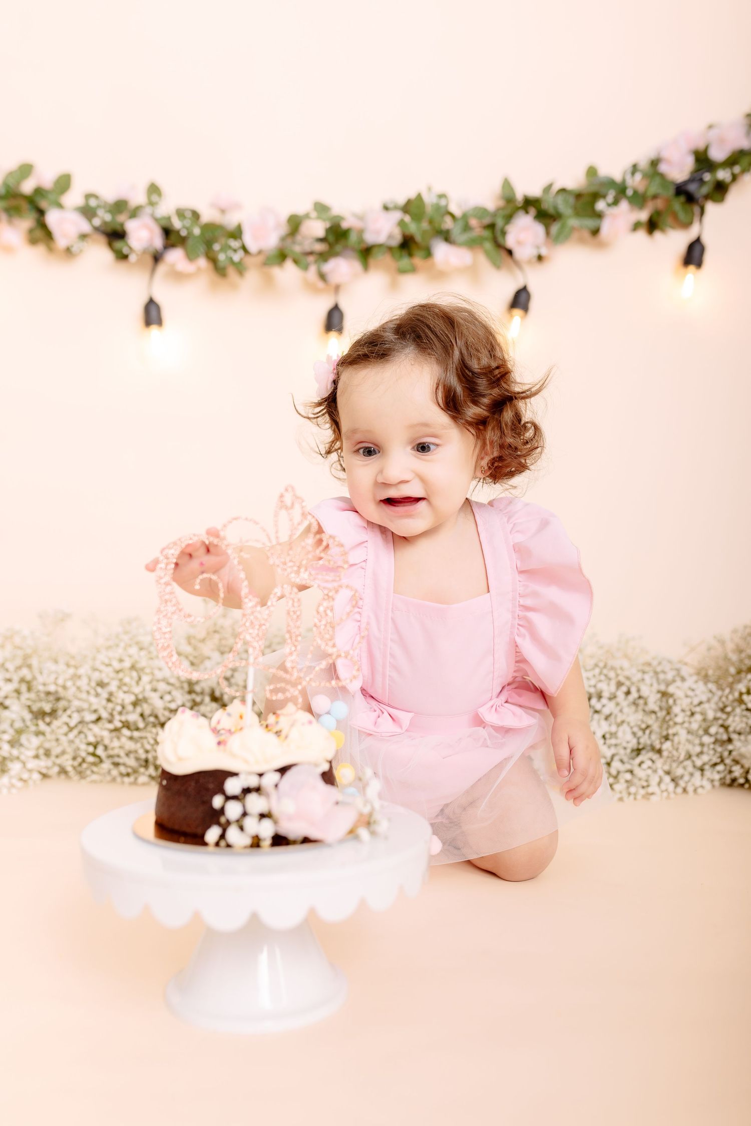 Smiling baby girl in pink outfit reaching for her first birthday cake on white stand with floral garland backdrop.