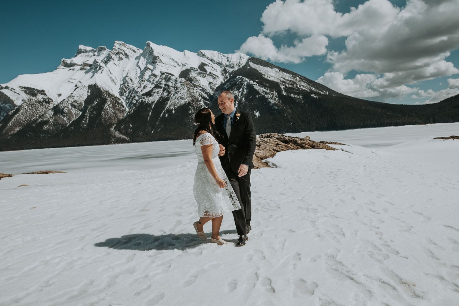 Couple in wedding attire holding hands on snowy landscape with snow-capped mountains in background.