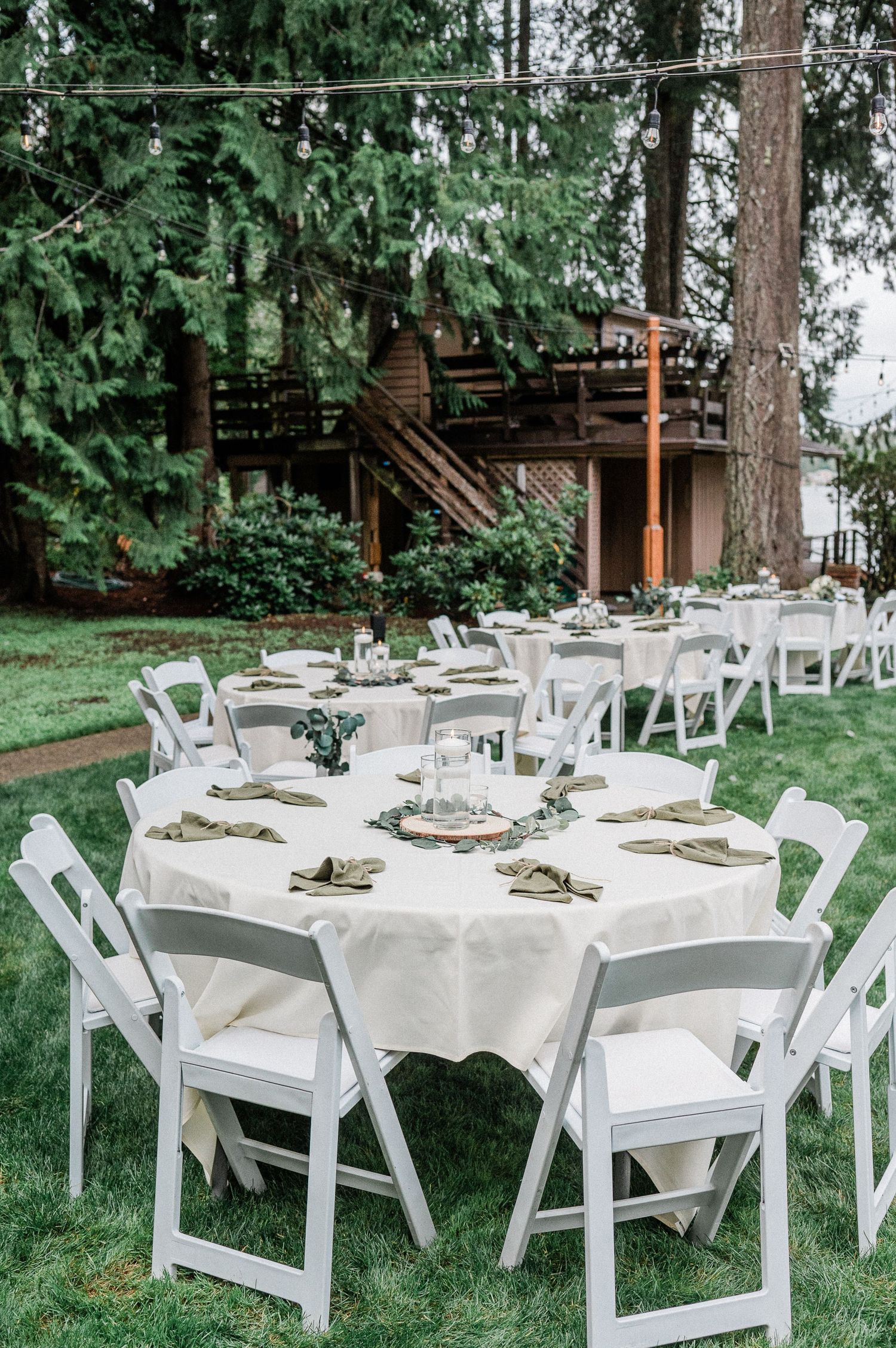 Elegant outdoor wedding reception with white folding chairs and round tables set on green lawn surrounded by tall pine trees.