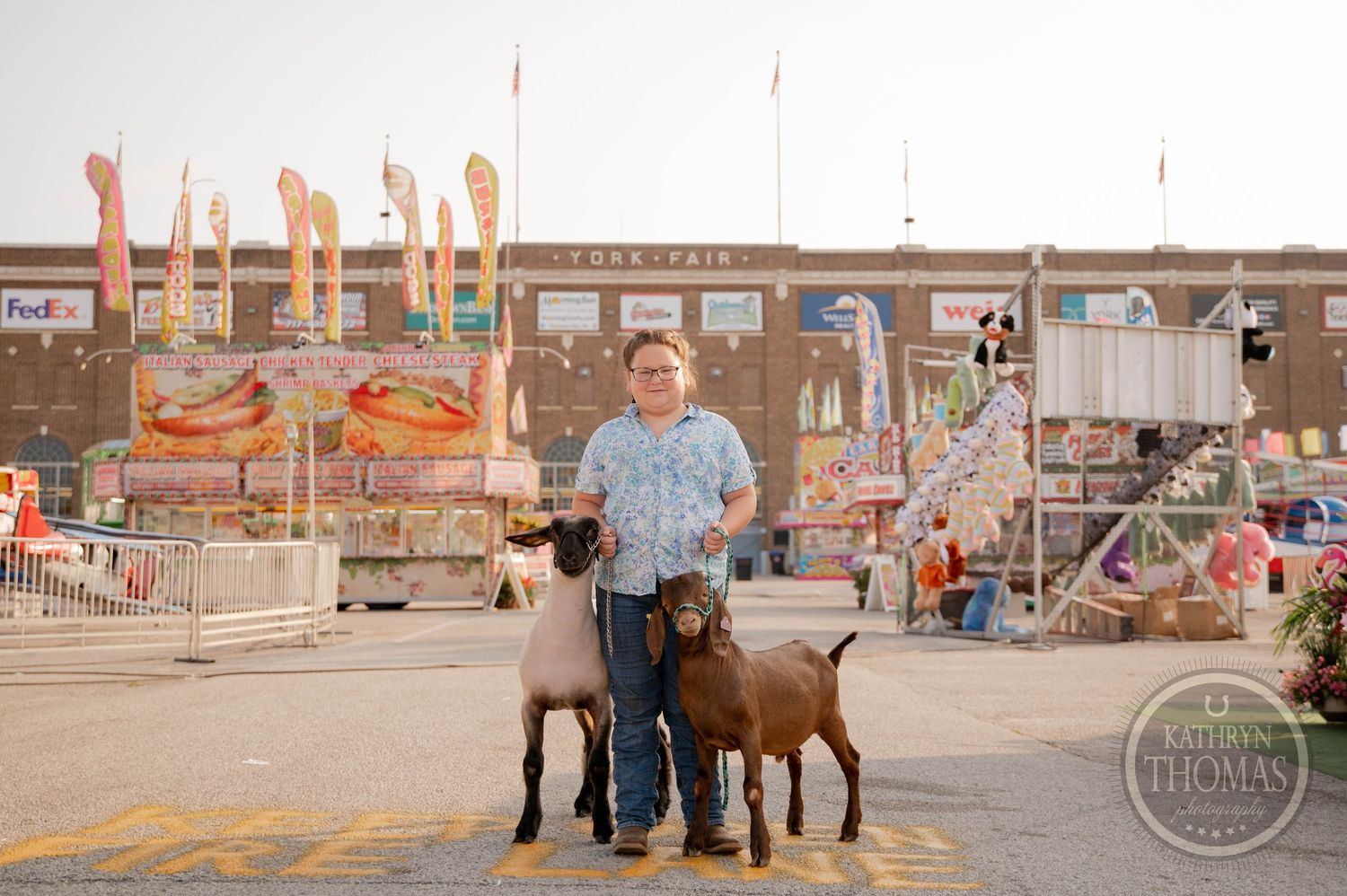 A boy stands with two brown goats at a county fair, with colorful carnival rides and food stands in the background.