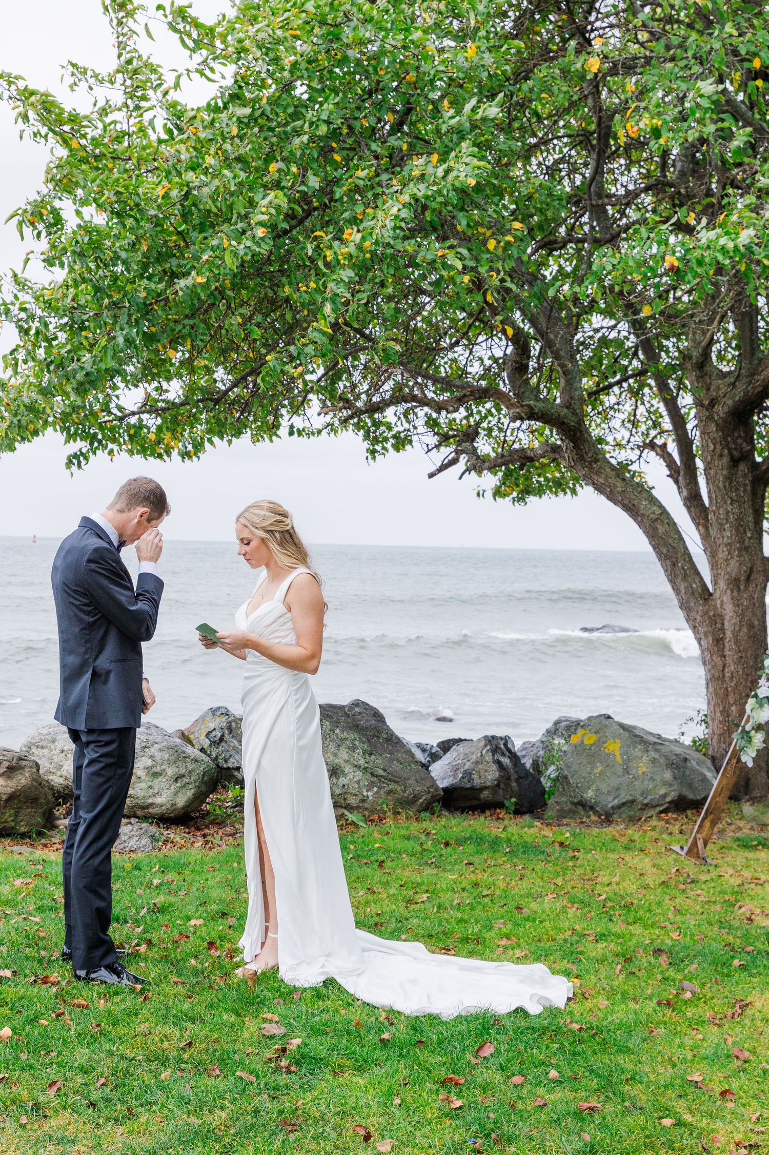 Bride and groom share a romantic kiss outdoors by the water during their wedding ceremony with bridal party watching.