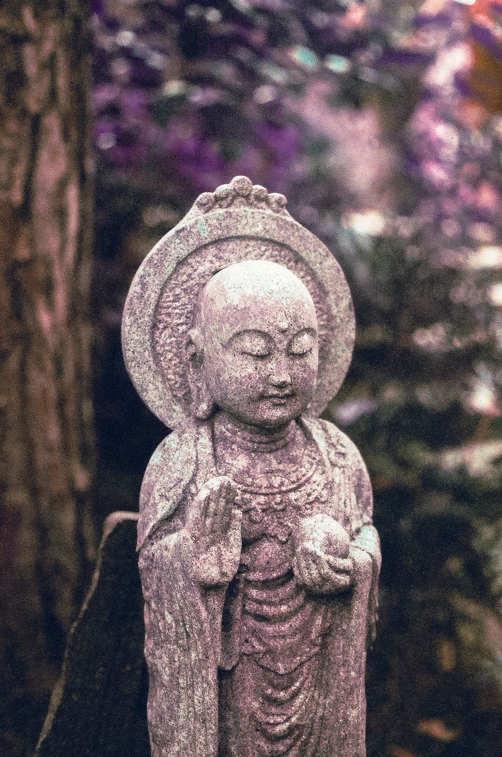 Stone Buddhist statue with halo, hands in prayer, surrounded by purple blossoms and trees in a serene garden setting.