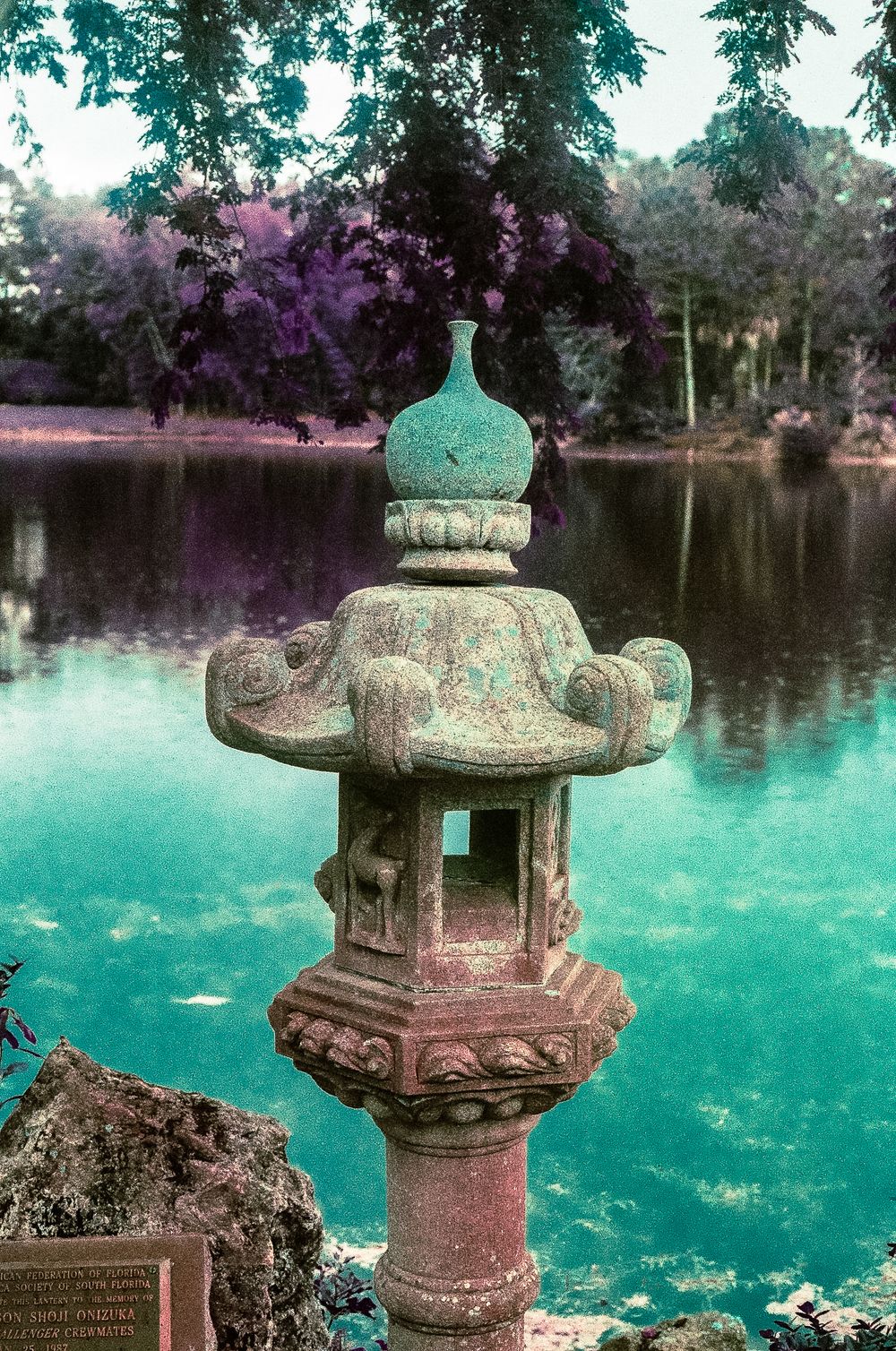 Stone Japanese lantern sculpture beside a vibrant turquoise pond surrounded by lush purple and green trees.