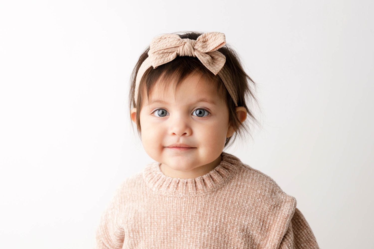 Adorable baby girl with blue eyes wearing a beige knit sweater and bow headband, smiling against a white background.