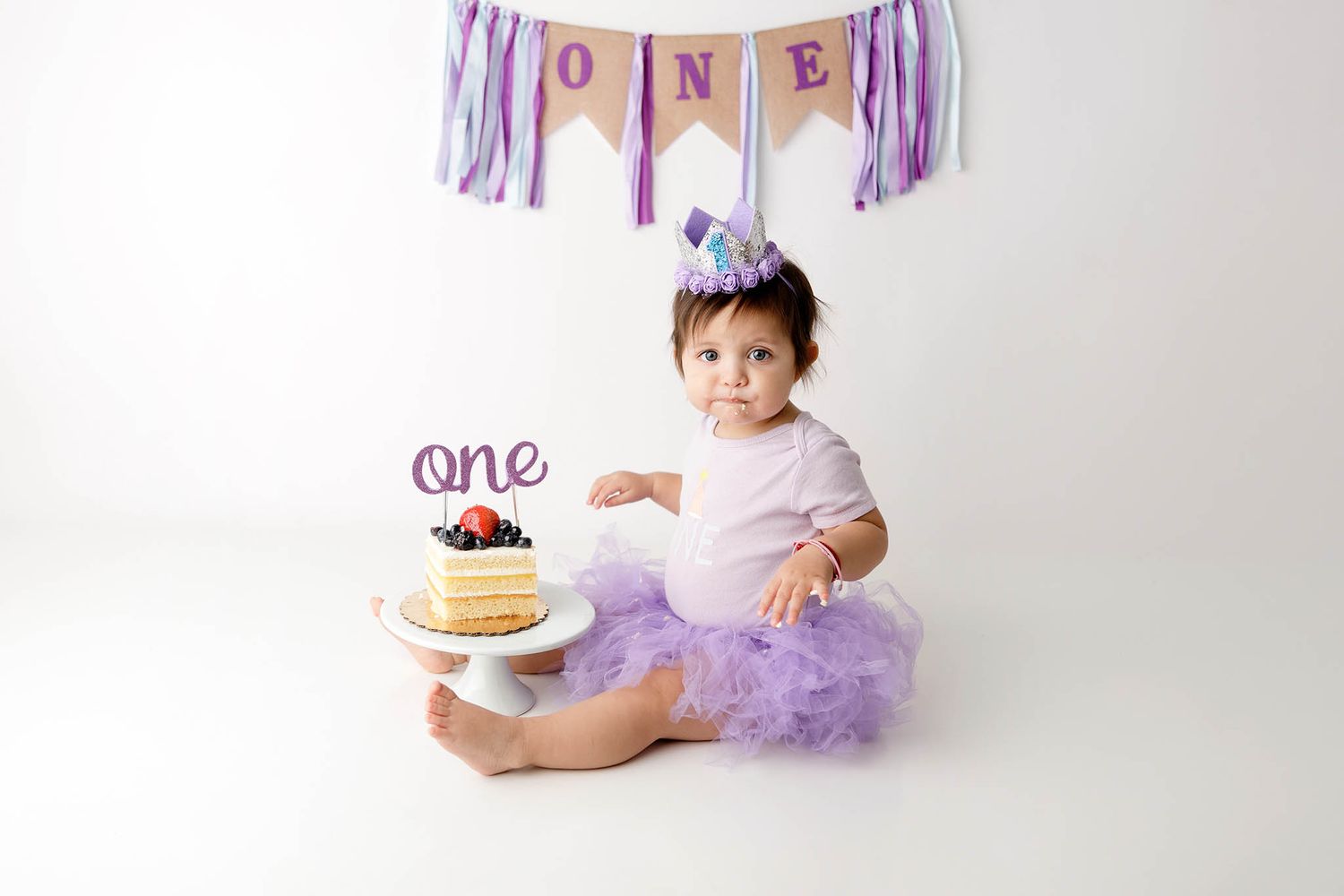 Baby girl in purple tutu sits beside a berry cake celebrating her first birthday with ONE banner backdrop.
