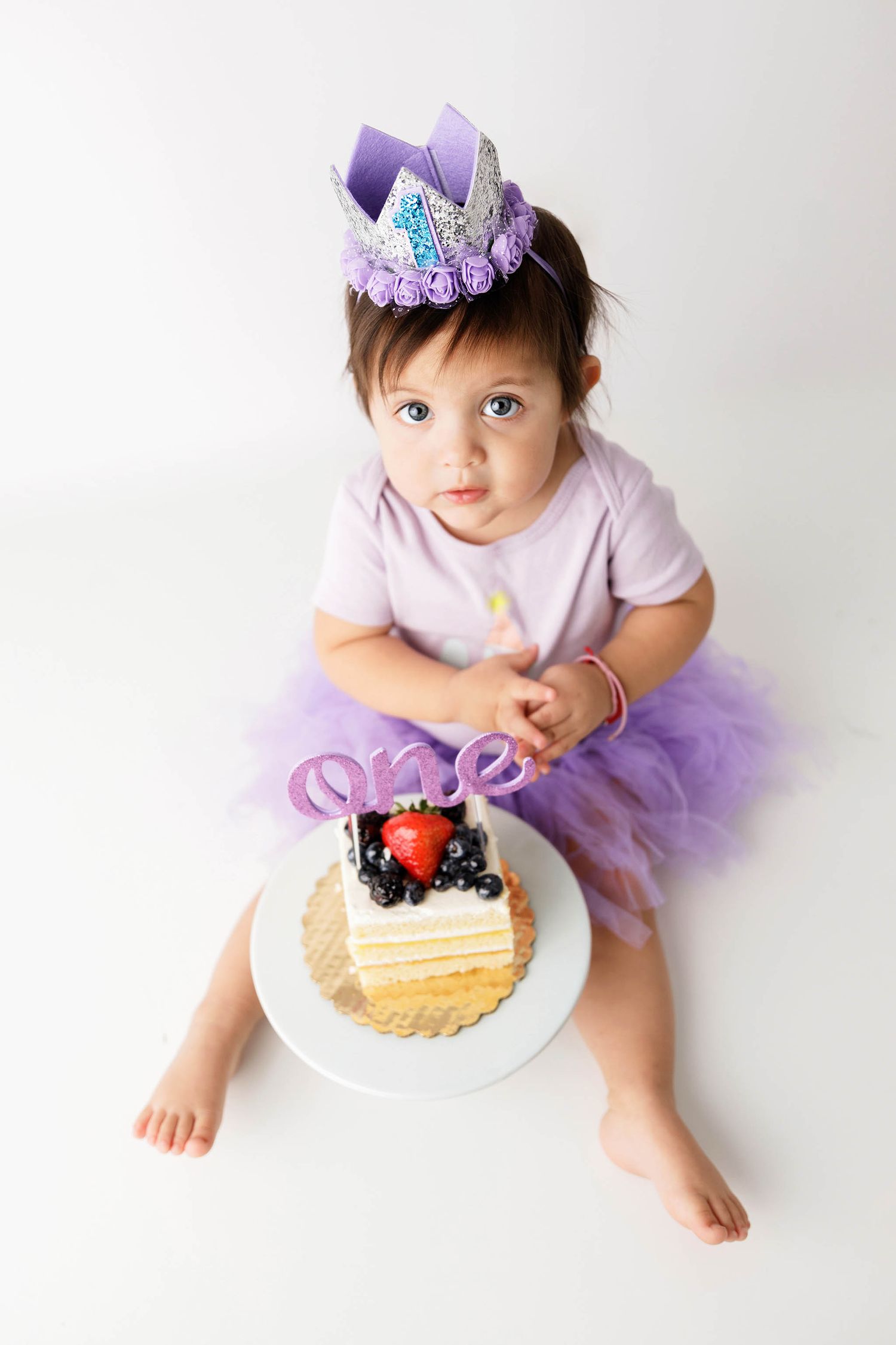 Baby girl in purple tutu and crown sits beside a first birthday cake decorated with fresh berries.