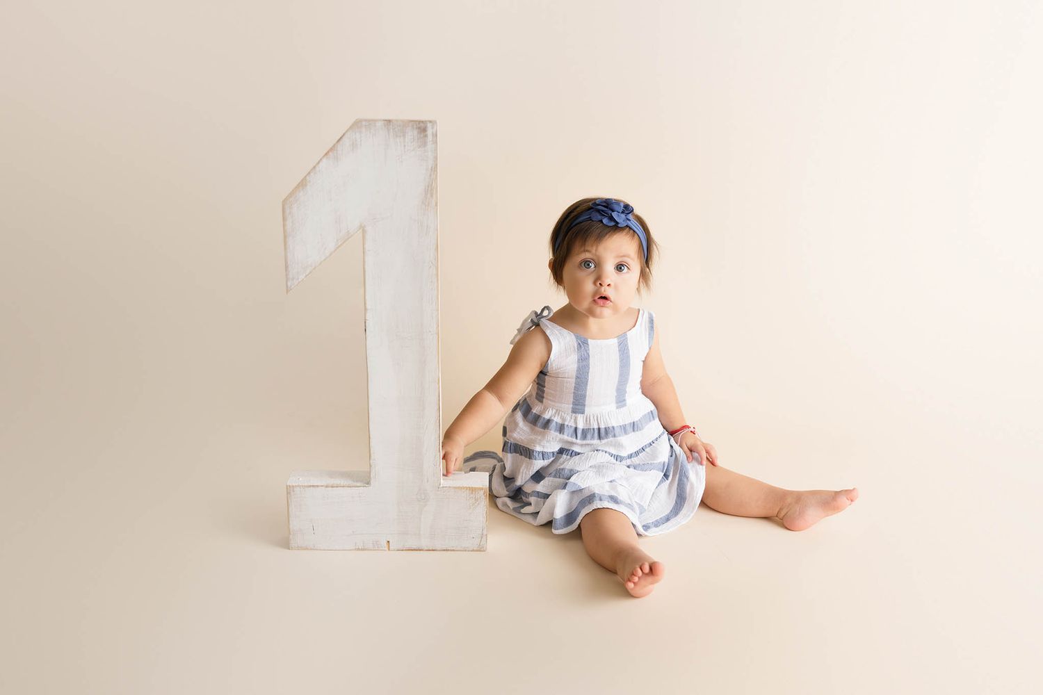 Baby girl in striped romper sits beside a large number one prop, celebrating her first birthday milestone.