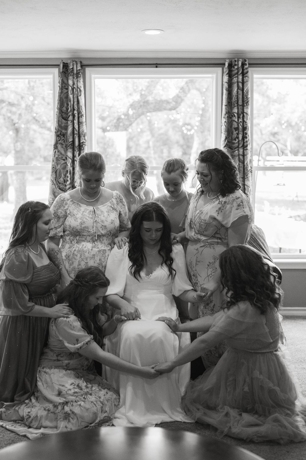 Emotional black and white wedding photography of a bride sitting in a chair surrounded by bridesmaids holding hands and praying