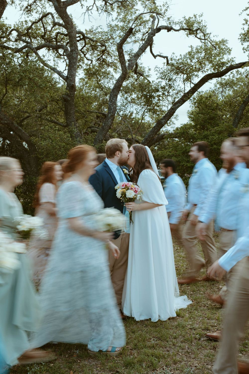 Creative wedding photography of a bride and groom kissing outdoors in Wimberley, TX, with bridesmaids and groomsmen walking past in motion b