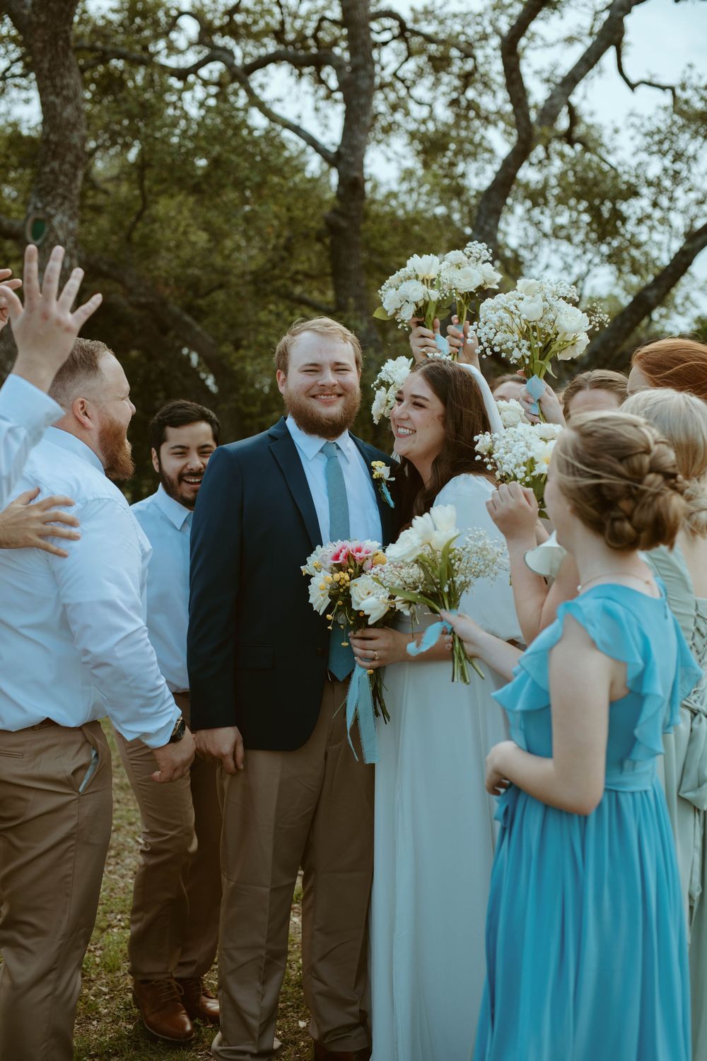 A happy bride and groom smiling while surrounded by their wedding party in a joyful group hug under oak trees in Wimberley, Texas.