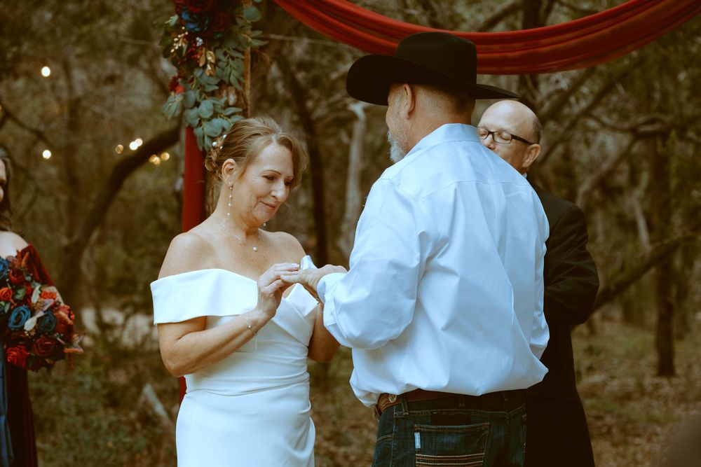 A groom wearing a cowboy hat places a ring on his bride's finger during an outdoor Texas Hill Country wedding ceremony in Wimberley, TX.