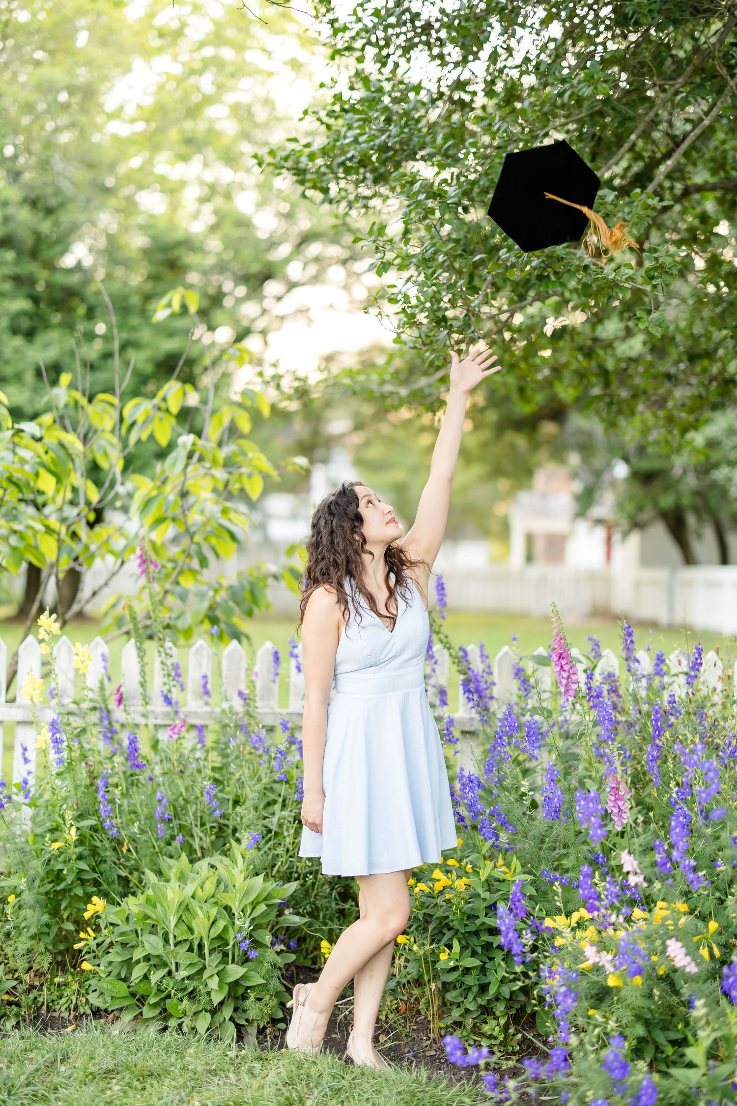 Senior photography of a recent graduate of the William and Mary Law School throwing her cap in a flower-filled garden in Williamsburg.