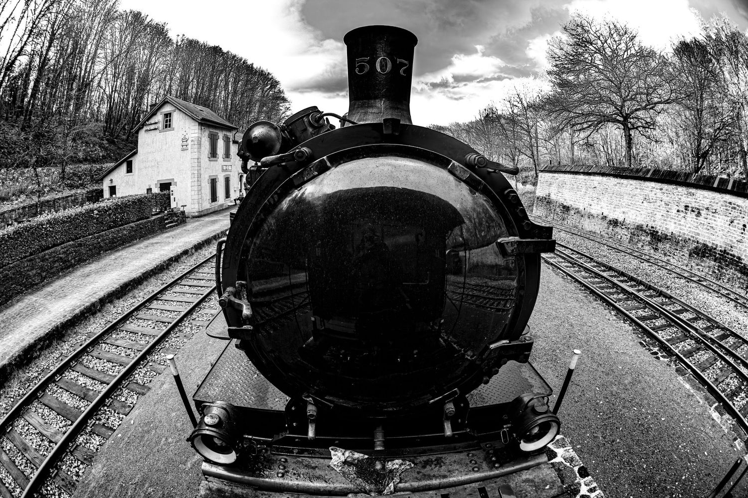 Black and white fisheye close-up of a vintage steam locomotive facing forward on railway tracks near a station.