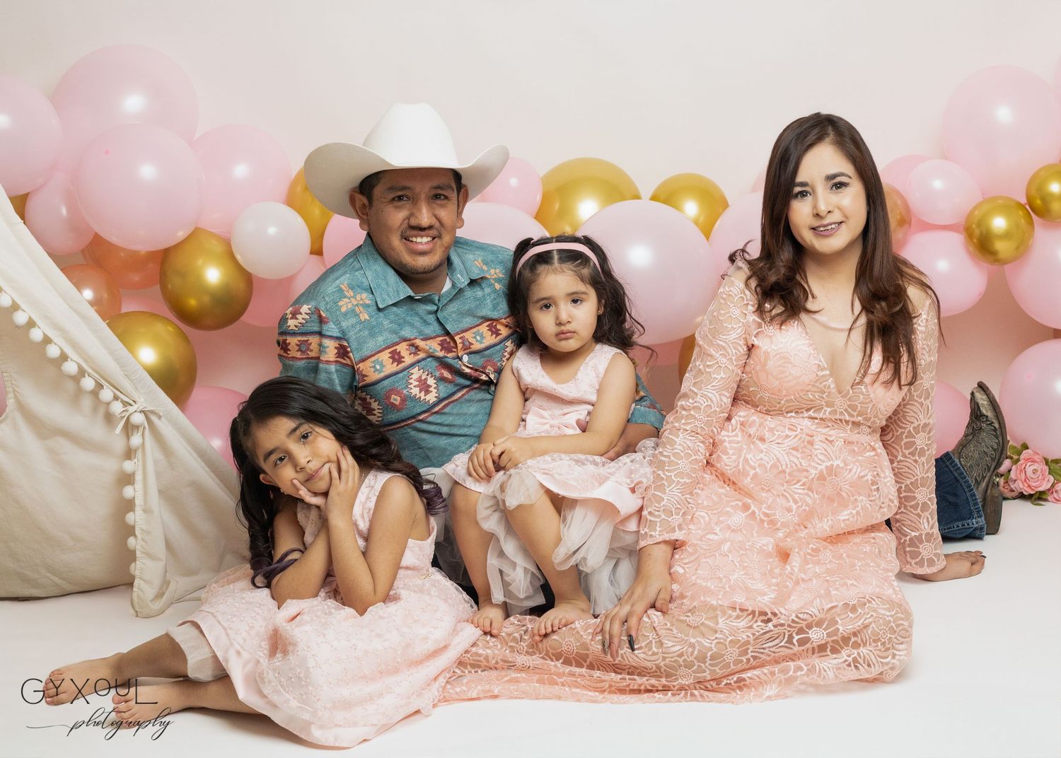 Family of four posing with pink and gold balloons, wearing elegant outfits, celebrating a special occasion in a studio.